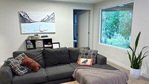 Un salon moderne avec un canapé gris, des coussins à motifs, une table basse en bois et une grande fenêtre donnant sur un paysage verdoyant.