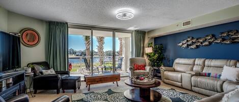 Amplia sala de estar con un sofá grande, una mesa de centro y un televisor. Hay un balcón con vistas a palmeras y una piscina.