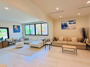 Ein großzügiges Wohnzimmer mit einem weißen Sofa, einem beigen Sofa, einem Couchtisch und einem großen Fenster mit Blick auf Grünflächen.