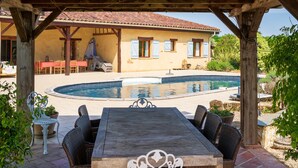 Un espacio exterior cubierto con piscina, mobiliario de mimbre y un edificio con ventanas y contraventanas.