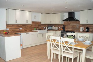 A modern kitchen with white cabinets, a wooden backsplash, and a dining area with white chairs and a wooden table. - Willoughby Cottage - Rustic Retreat in Crieff (Muthill)