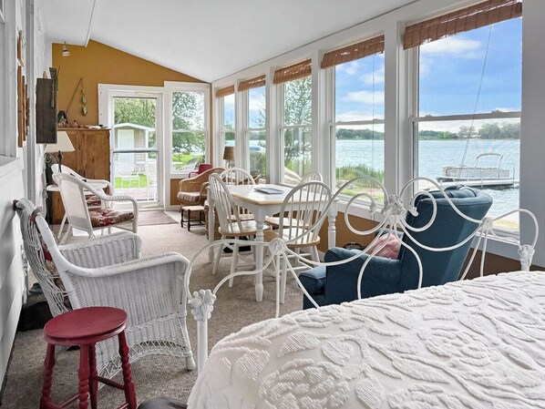 A room with a bed, white wicker chairs, a dining table, and large windows overlooking a body of water.