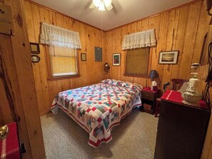 Une pièce aux murs en bois, un lit recouvert d’une courtepointe, deux fenêtres avec des rideaux, une table de chevet avec une lampe et une commode recouverte d’un tissu rouge.