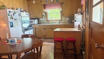 A compact kitchen with wooden cabinets, a dining table with chairs, and a window with a view of greenery.