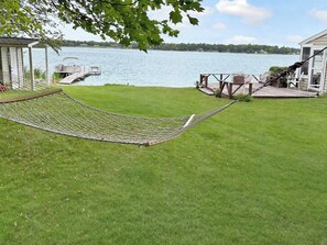 A hammock on a grassy lawn with a dock and chairs, overlooking a body of water.