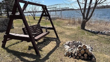 A wooden swing set and a pile of firewood by a lakeside.