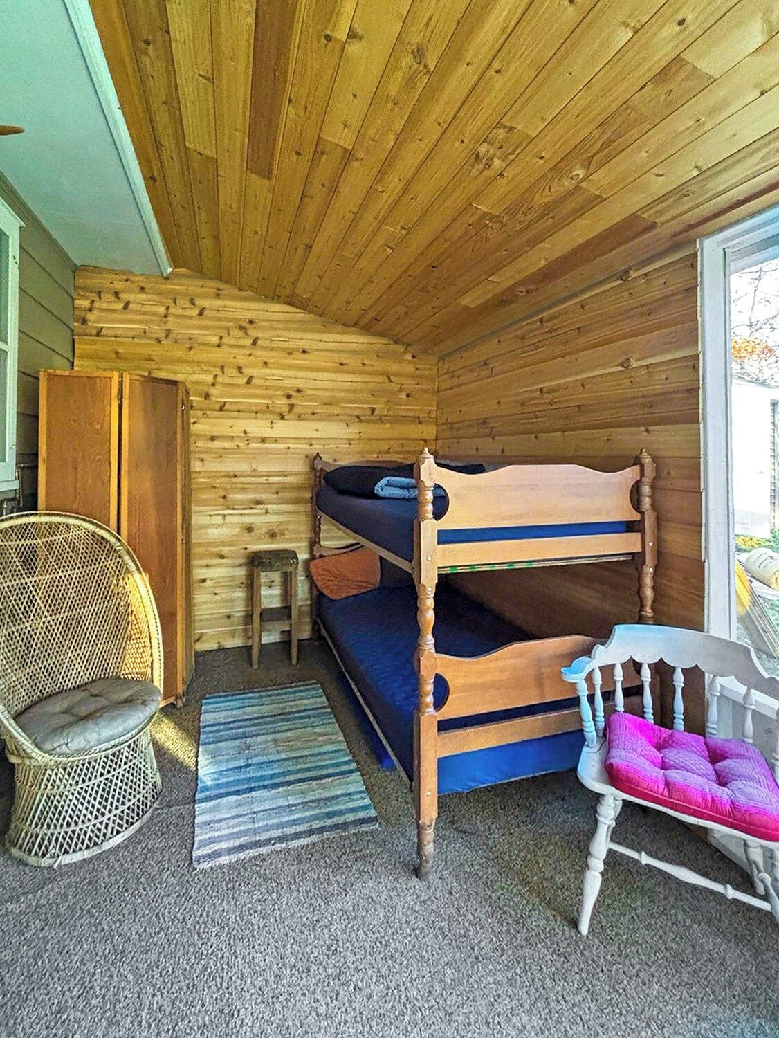 A small, cozy room with a bunk bed, a wicker chair, a small table, and a window.