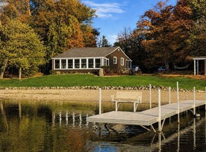 Une maison avec un quai s’avançant dans une étendue d’eau calme, entourée d’arbres aux feuilles d’automne.