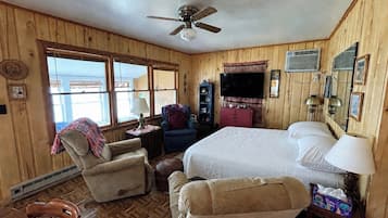 A wooden-paneled room with a bed, two armchairs, a television, and a ceiling fan.