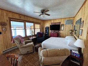 A wooden-paneled room with a bed, two armchairs, a television, and a ceiling fan.
