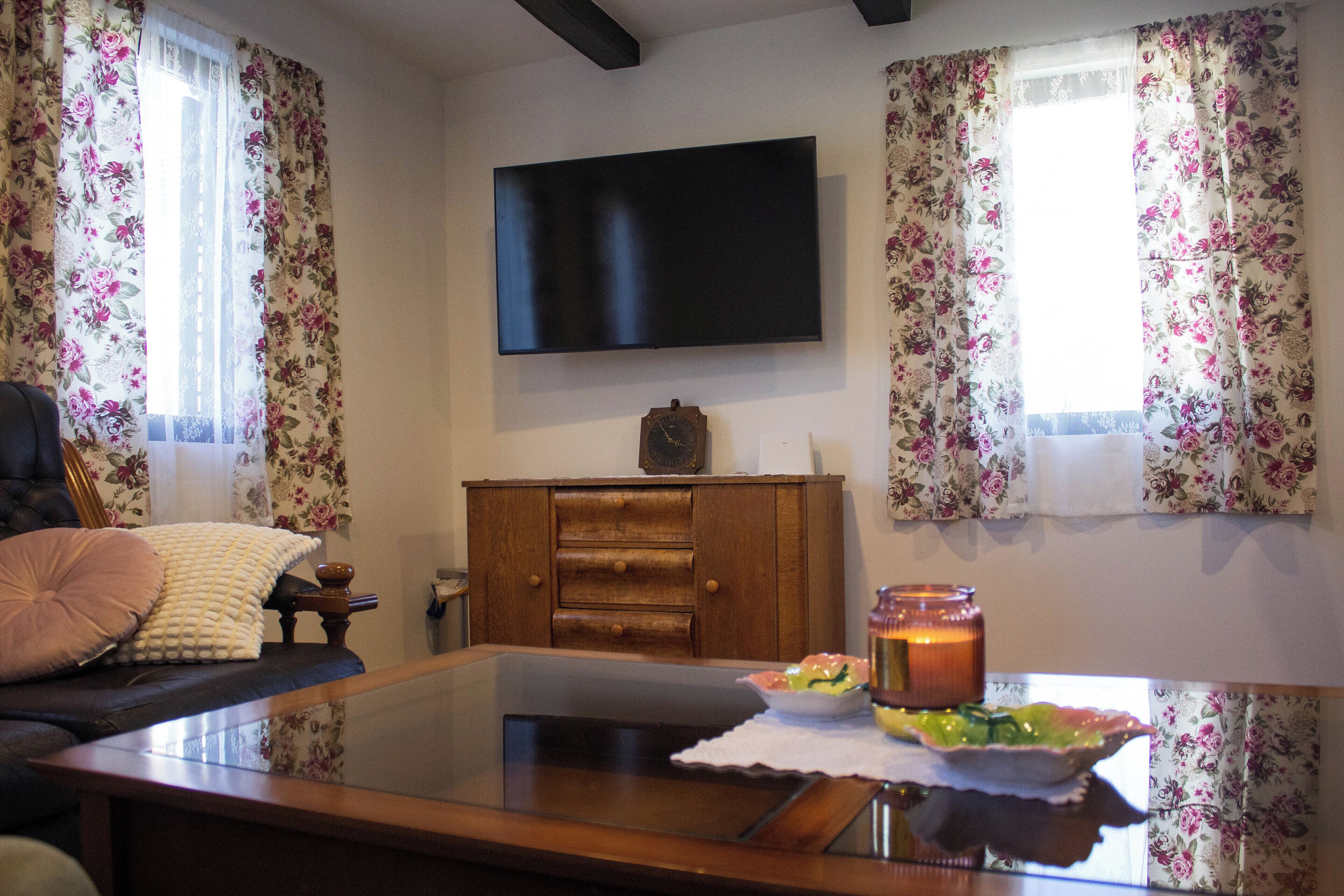 A living room with a flat-screen TV mounted on the wall, a wooden cabinet, a glass coffee table, and floral curtains.
