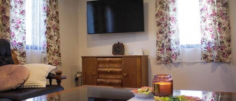 A living room with a flat-screen TV mounted on the wall, a wooden cabinet, a glass coffee table, and floral curtains.