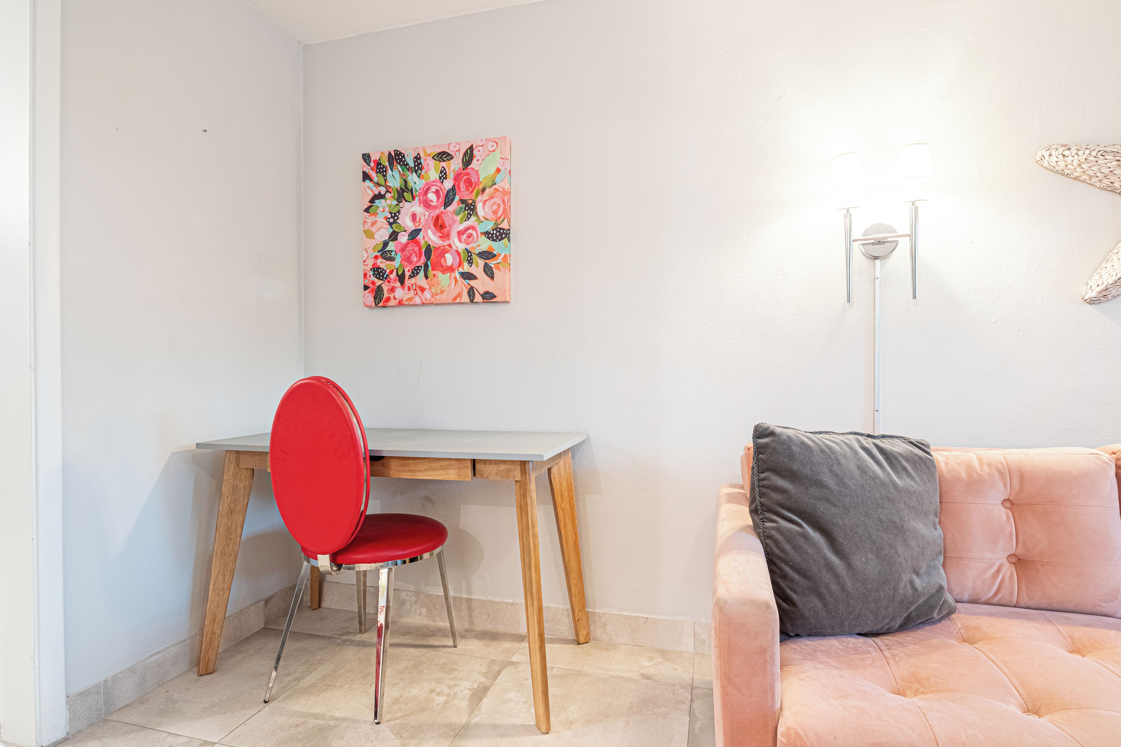 Une pièce avec un bureau en bois, une chaise rouge, un canapé rose avec un coussin gris et un tableau représentant des motifs floraux accroché au mur.