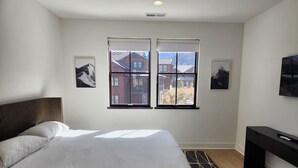 A bedroom with a large bed, a desk, and two windows with a view of buildings. - Snowcreek Resort Vacation Rental 1543 (Mammoth Lakes)