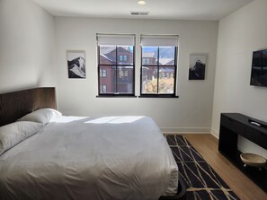 Une chambre à coucher avec un grand lit, un bureau et deux fenêtres donnant sur des immeubles.