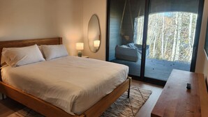 A bedroom with a bed, bedside lamps, a mirror, a wooden nightstand, a small table, and a sliding glass door leading to a balcony with a hanging chair. - Snowcreek Resort Vacation Rental 1543 (Mammoth Lakes)