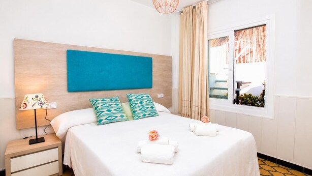 A hotel room with a bed, a bedside table with a lamp, a window with curtains, and a view of the outside. - Villa Maria de Bonaire (Mallorca)