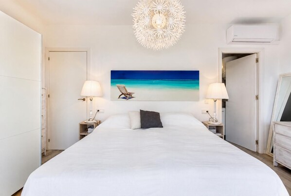 Een hotelkamer met een groot bed, twee nachtkastjes met lampen, een plafondlamp en een schilderij met strandthema aan de muur.