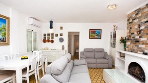 A modern living room with a grey sofa, a dining table with chairs, a fireplace, and a kitchen area with a white countertop and cabinets. - Villa Maria de Bonaire (Mallorca)