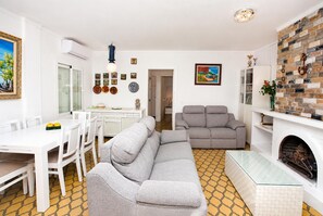 A modern living room with a grey sofa, a dining table with chairs, a fireplace, and a kitchen area with a white countertop and cabinets. - Villa Maria de Bonaire (Mallorca)