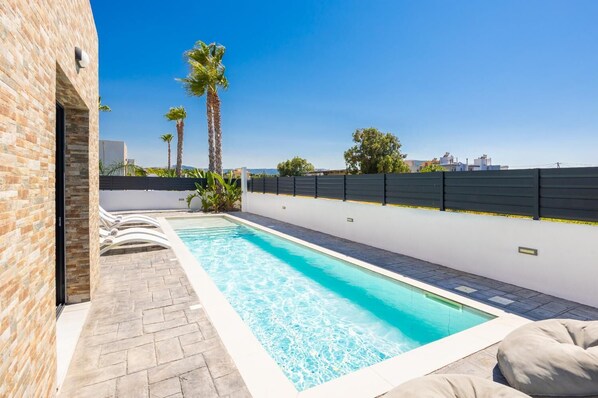 A swimming pool with a brick wall on one side and a white wall on the other, surrounded by palm trees and a fence. - Villa Salty Sun 5 - Two Bedroom Villa, Sleeps 4 (Kremasti)