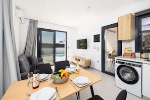 A modern kitchen with a washing machine, dining area with table set for two, and a view of the pool. - Villa Salty Sun 5 - Two Bedroom Villa, Sleeps 4 (Kremasti)