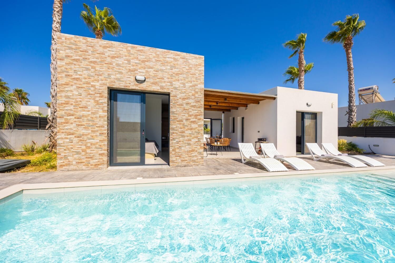 A modern poolside area with a stone building, a covered seating area, and lounge chairs.