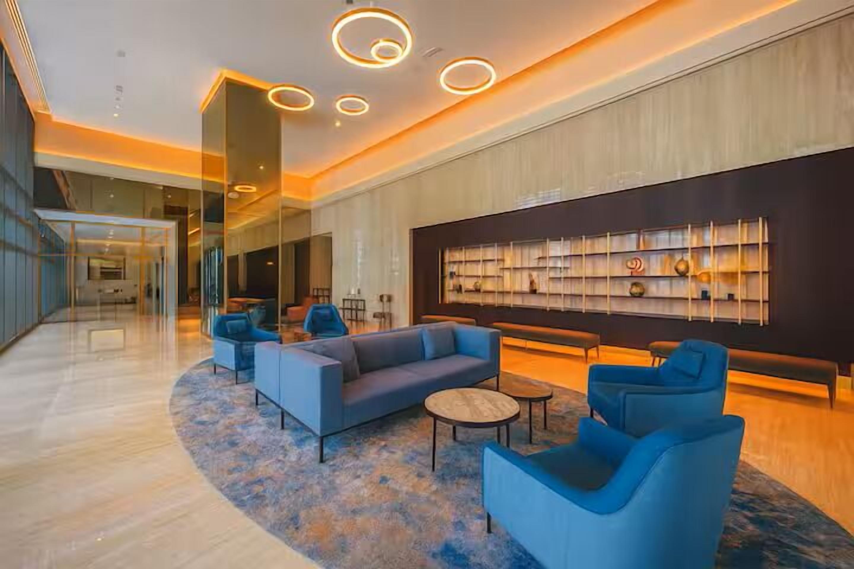 A modern lobby with a blue sofa set, round coffee table, and blue armchairs. There is a large bookshelf with decorative items on the wall.