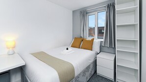 Ein Hotelzimmer mit einem ordentlich bezogenen Bett, einem Nachttisch mit Lampe, einem Fenster mit Vorhängen und integrierten Regalen.