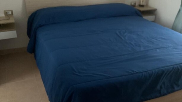 Un llit amb capçalera de fusta i estriat de color blau en una habitació amb terra de rajoles.