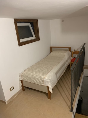A small, single-bed room with a wooden headboard, a window with a wooden frame, and a tiled floor.