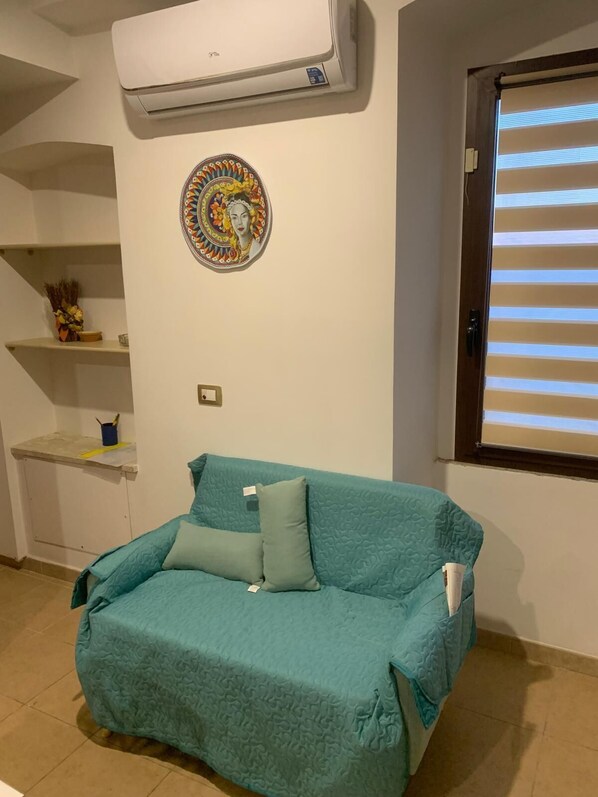 A teal-colored sofa with a patterned cover, a wall-mounted air conditioner, a decorative circular artwork, and a window with blinds.