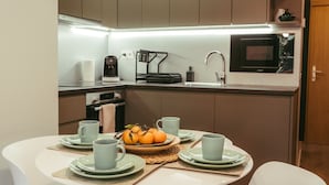 Een kleine keuken met moderne apparatuur, een eettafel gedekt met borden en kopjes, en een fruitschaal op het aanrecht.
