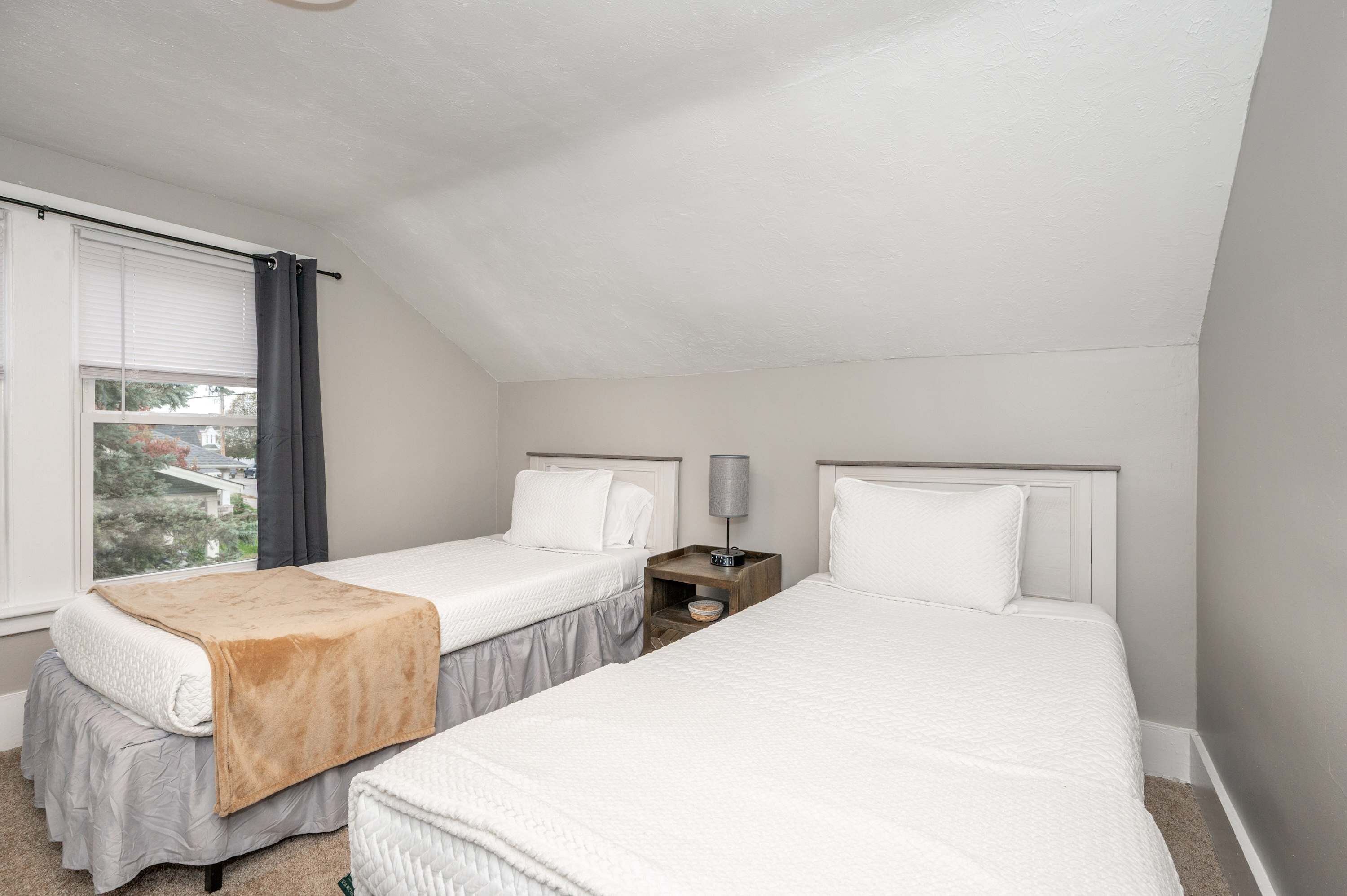 A bedroom with two beds, a window with curtains, a bedside table with a lamp, and a view of the outside.
