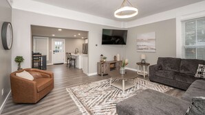 Ein modernes Wohnzimmer mit einem braunen Ledersofa, einem grauen Couchsofa und einem Couchtisch. An der Wand ist ein Fernseher angebracht, und im Hintergrund ist eine Küchenzeile erkennbar.