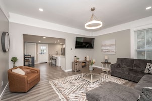 Uma sala de estar moderna com um sofá de couro marrom, um sofá cinza e uma mesa de centro. Há uma TV na parede e uma cozinha visível ao fundo.
