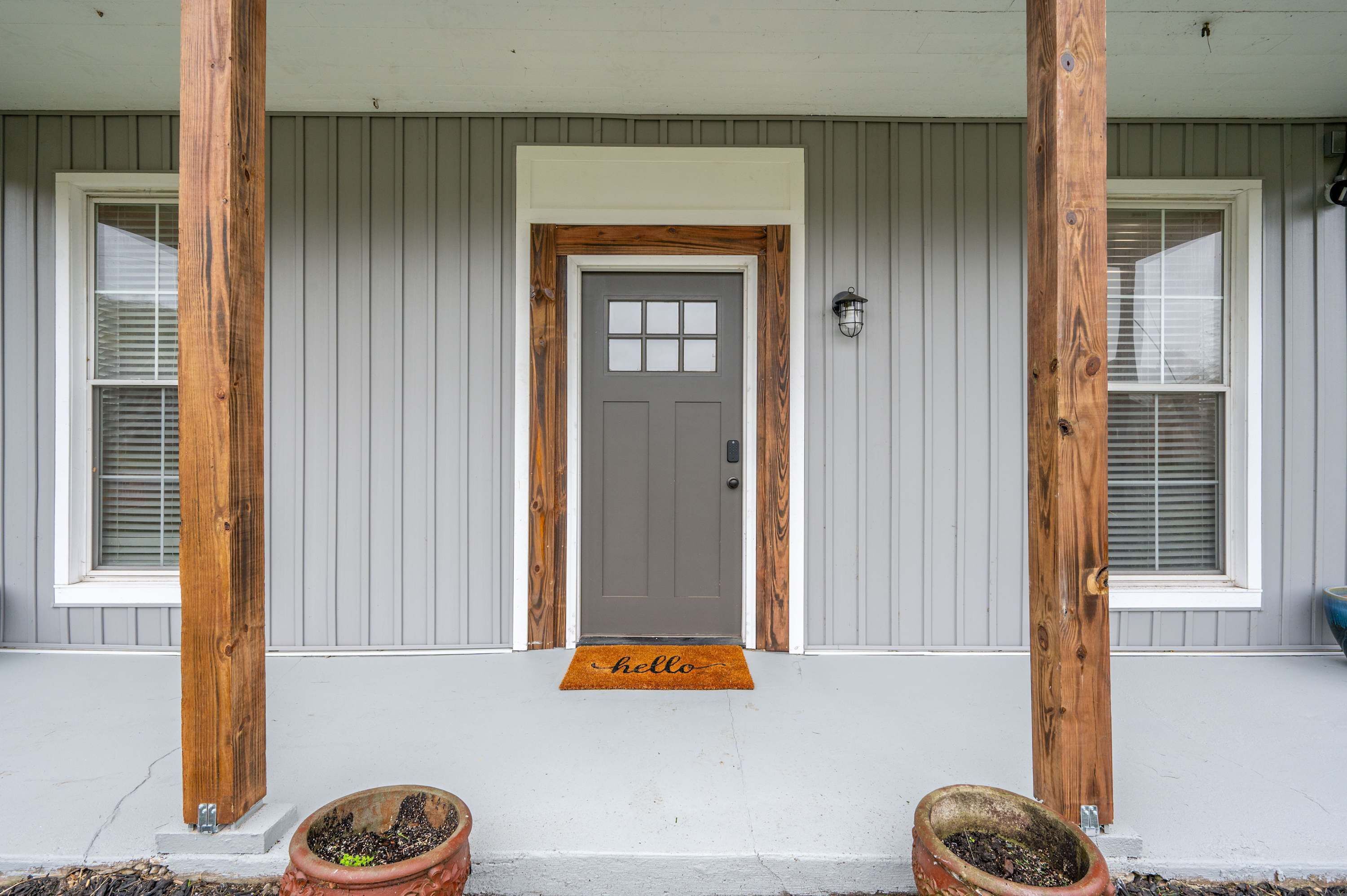 Una casa con fachada gris, una puerta principal gris y un porche de madera. Hay dos macetas con plantas en el porche.