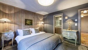 A bedroom with a wooden panel wall, a bed with a grey bedspread, two bedside lamps, a nightstand with a lamp, and a mirror. - MONTCLAIR HOME - Four Bedroom Apartment, Sleeps 8 (Verbier)