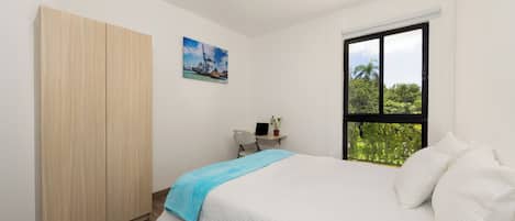 Un dormitorio con cama, armario, escritorio con silla, ventana con vistas a follaje y un cuadro en la pared.