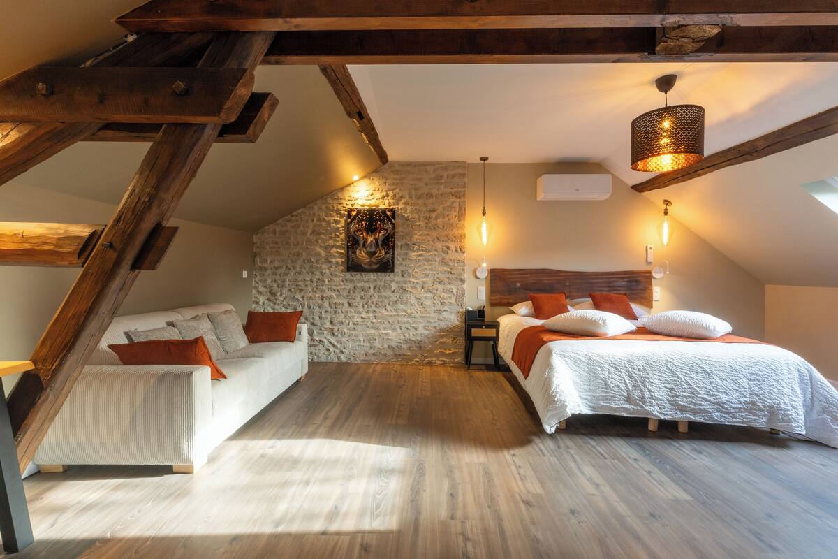 Quarto tipo loft aconchegante, com teto inclinado, vigas de madeira aparente, parede de pedra e uma cama grande com roupa de cama branca.