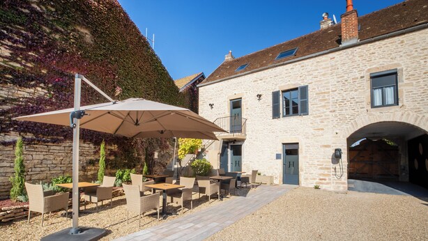 An outdoor patio with tables and chairs, a large umbrella, and a stone building with a door and window shutters. - Hotel les Vents d'Anges (Beaune)
