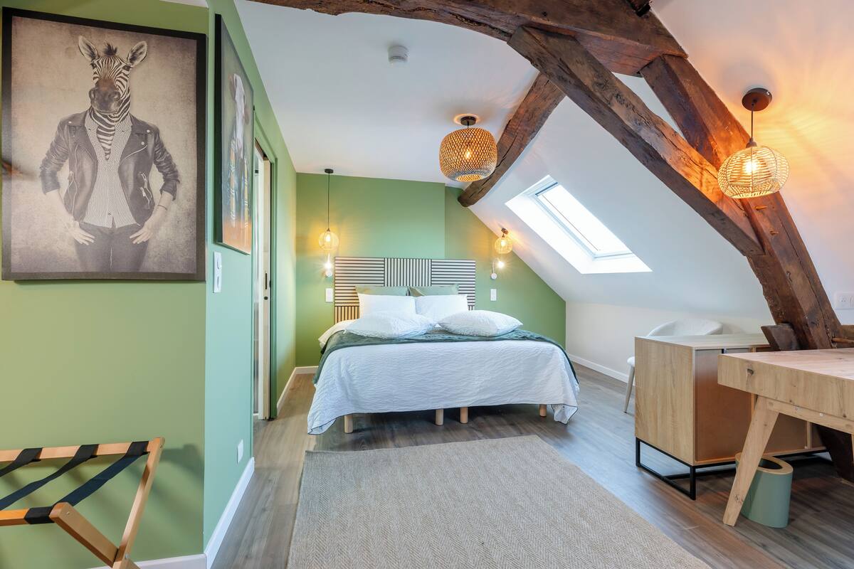 Um quarto de sótão acolhedor, com teto inclinado, cama com roupa de cama branca, uma secretária de madeira e uma grande obra de arte emoldurada na parede.