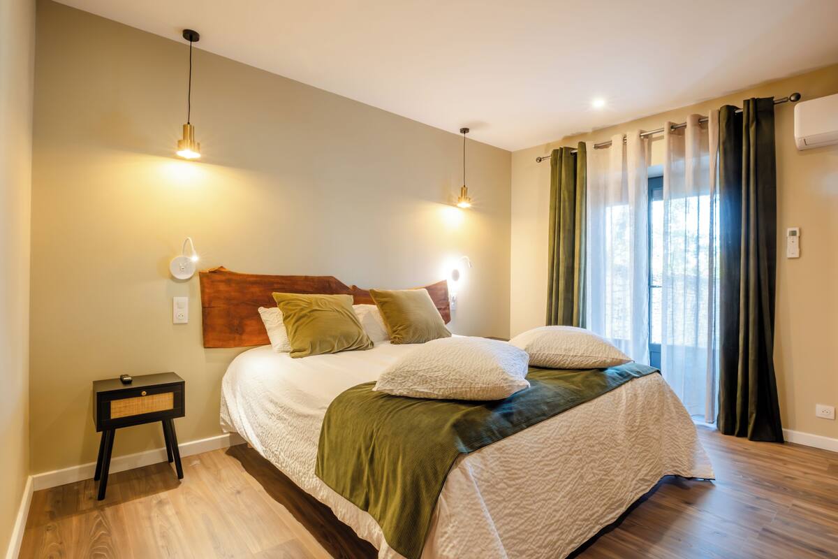 Quarto com cabeceira de madeira, cama com roupa de cama branca, duas luminárias de cabeceira, mesa de cabeceira e janela com cortinas.