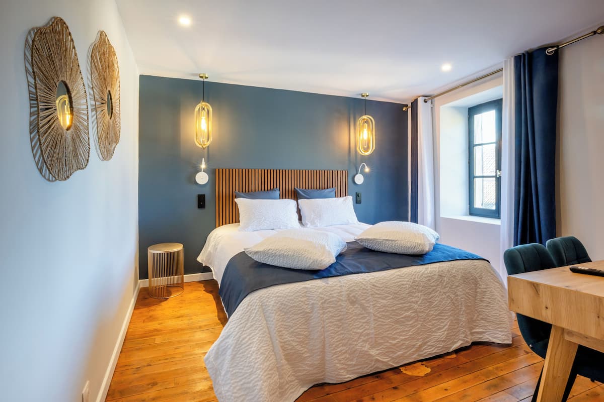 Quarto moderno com cabeceira de madeira, cama com roupa de cama branca e azul, duas arandelas de parede, uma mesa lateral redonda e uma janela com cortinas.