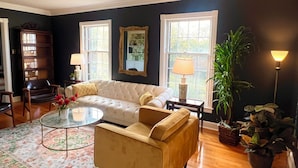A living room with a white sofa, a yellow armchair, a glass coffee table, and a floral rug. There are two windows with white frames, a mirror, and a lamp on a side table. - Massey's in the Park (Vicksburg)