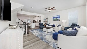 Ein modernes Wohnzimmer mit einem weißen Sofa, blauen Akzentkissen, einem Couchtisch und einem blauen Teppich. Ein Deckenventilator ist zu sehen, und eine Treppe führt auf eine obere Ebene.