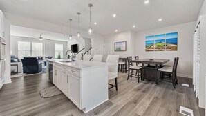 A modern kitchen with white cabinetry, a central island, and pendant lighting. There is a dining area with a dark wooden table and chairs. - Golfer's Paradise - 5-bedroom luxe townhome with golf simulator & pool (North Myrtle Beach)