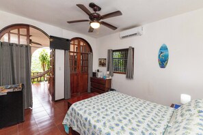 A bedroom with a bed, a ceiling fan, a window with curtains, a surfboard decoration, and a wooden door. - Villas el Parque #18B: Central Studio Apartment, Pool, AC, WiFi & Wildlife (Quepos)