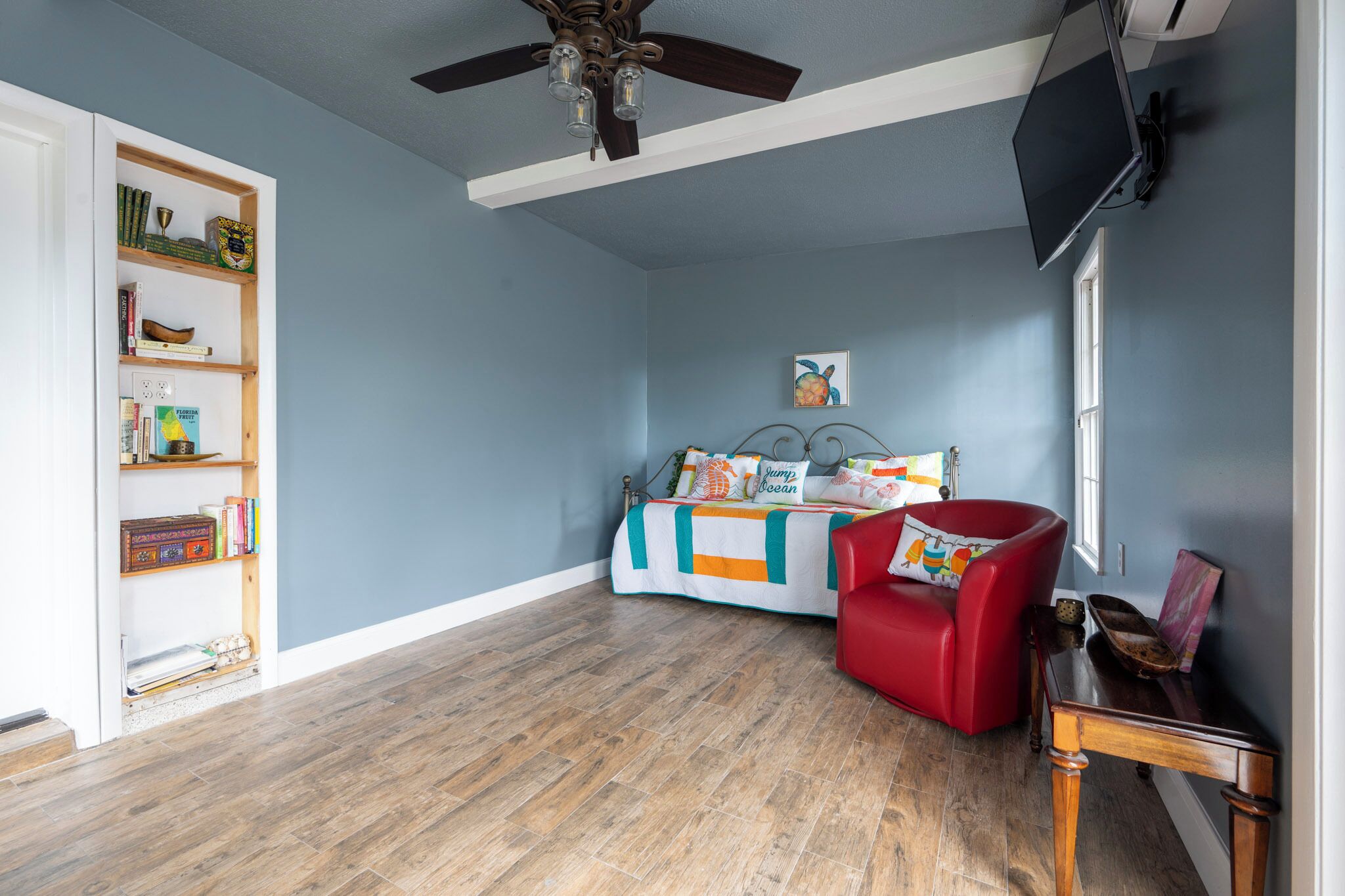 A room with a bed, a red chair, a bookshelf, and a ceiling fan.