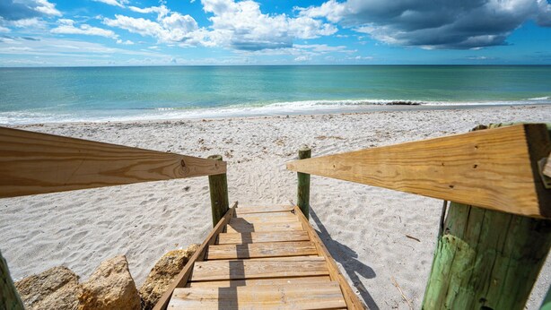 Un escalier en bois mène à une plage de sable, d’où l’on a une vue dégagée sur l’océan et un ciel partiellement nuageux.